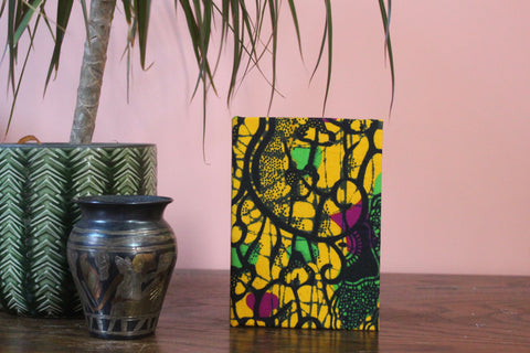 Cotton ankara fabric covered notebook. The book is yellow with pink, green and black abstract pattern overlayed.